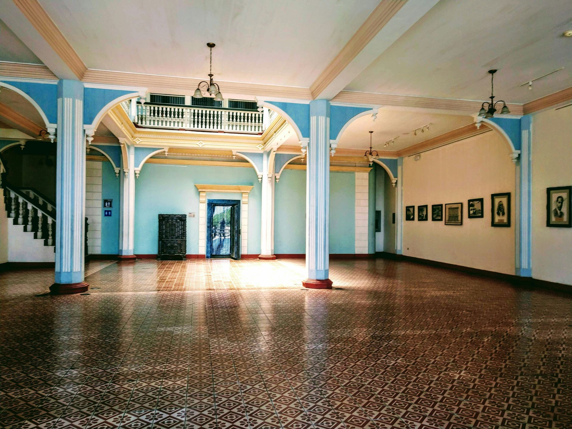 Spacious interior with tiled floor, blue walls, columns, balcony, artwork, and a grand staircase.