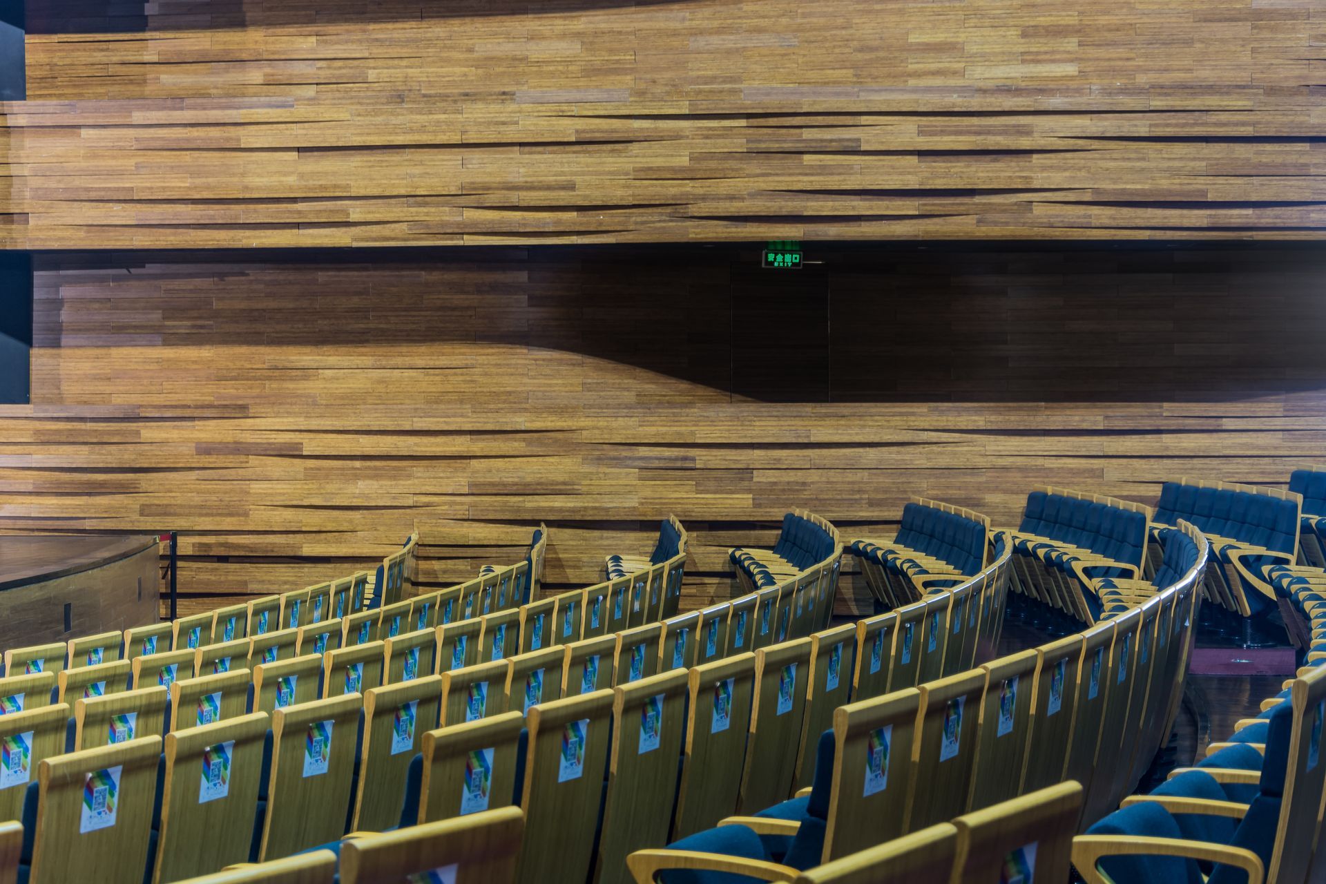 Auditorium interior: Rows of seats face a stage. Wood-paneled walls, an exit sign.