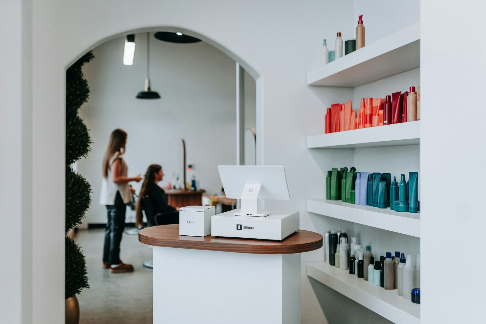 Salon interior: woman getting hair styled near desk with payment system and shelves of colorful products.