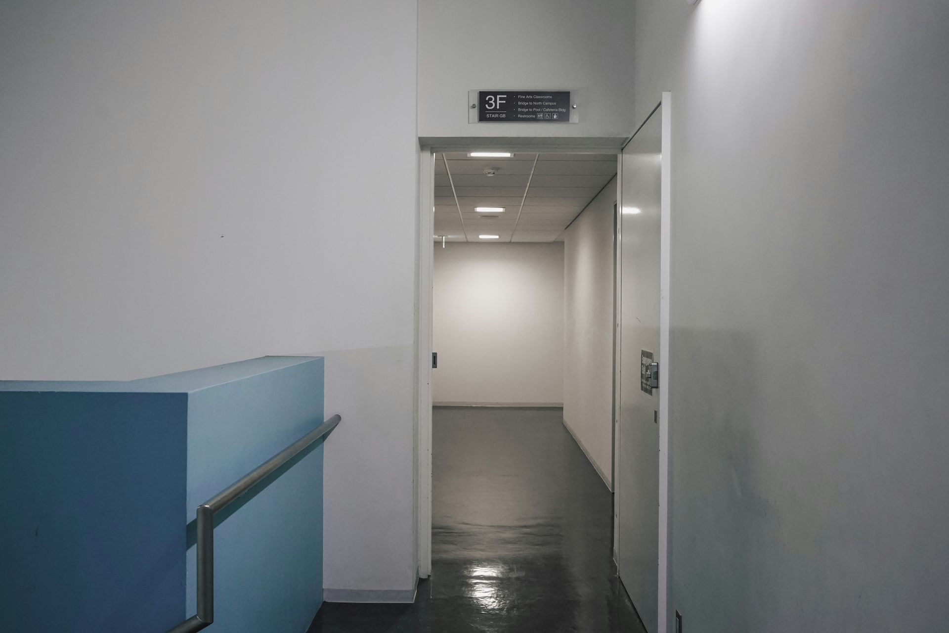 Hallway with white walls, a blue staircase, and an open door leading to a long, bright corridor.