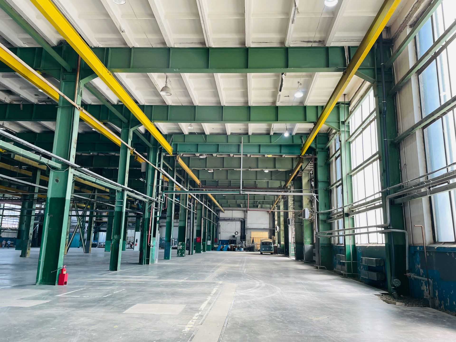 Empty industrial warehouse interior with green beams, yellow rails, and large windows.