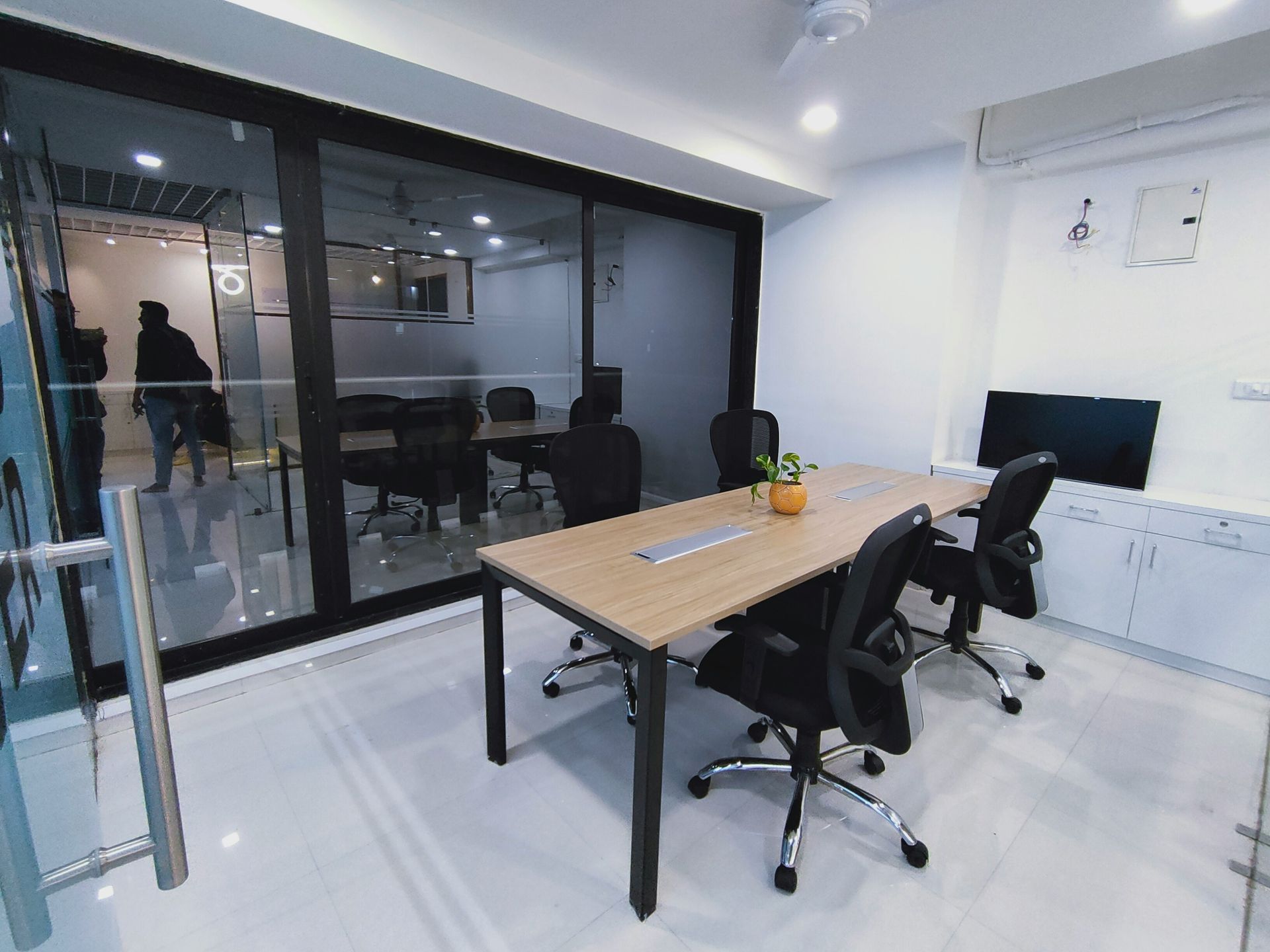 Conference room with long table, chairs, and glass walls. A person walks outside.