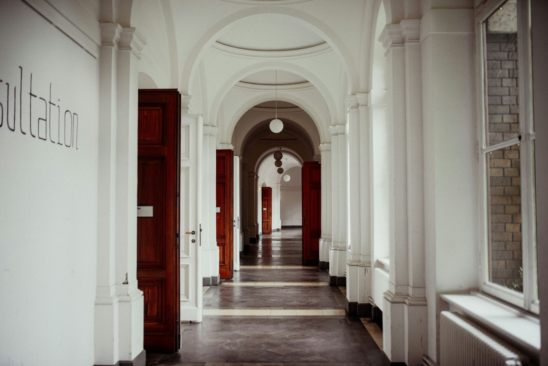 Long, white hallway with arched ceilings, columns, and open brown doors. Sunlight enters from a window.