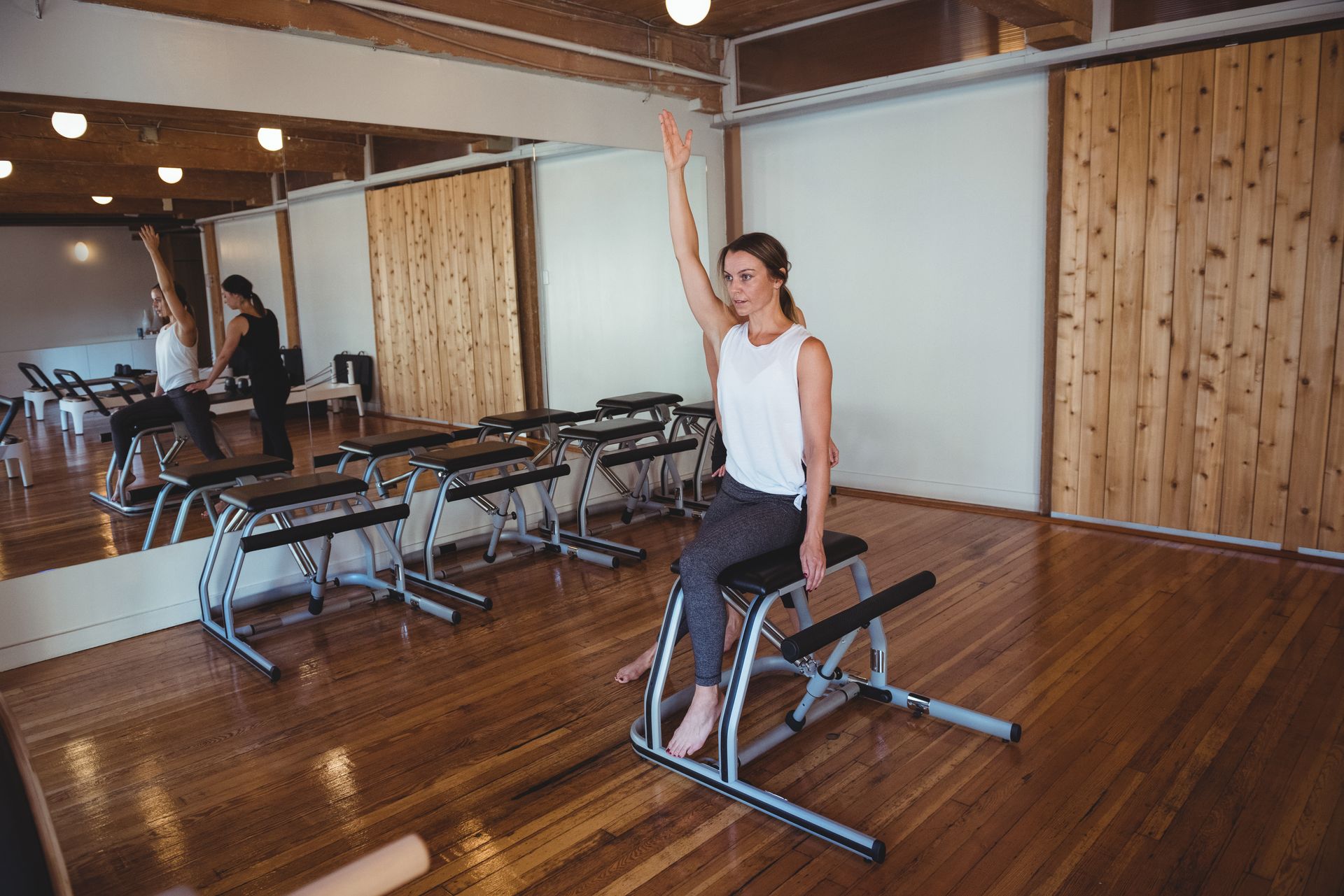 Woman in pilates class, seated, arm raised. Wooden floor and walls.