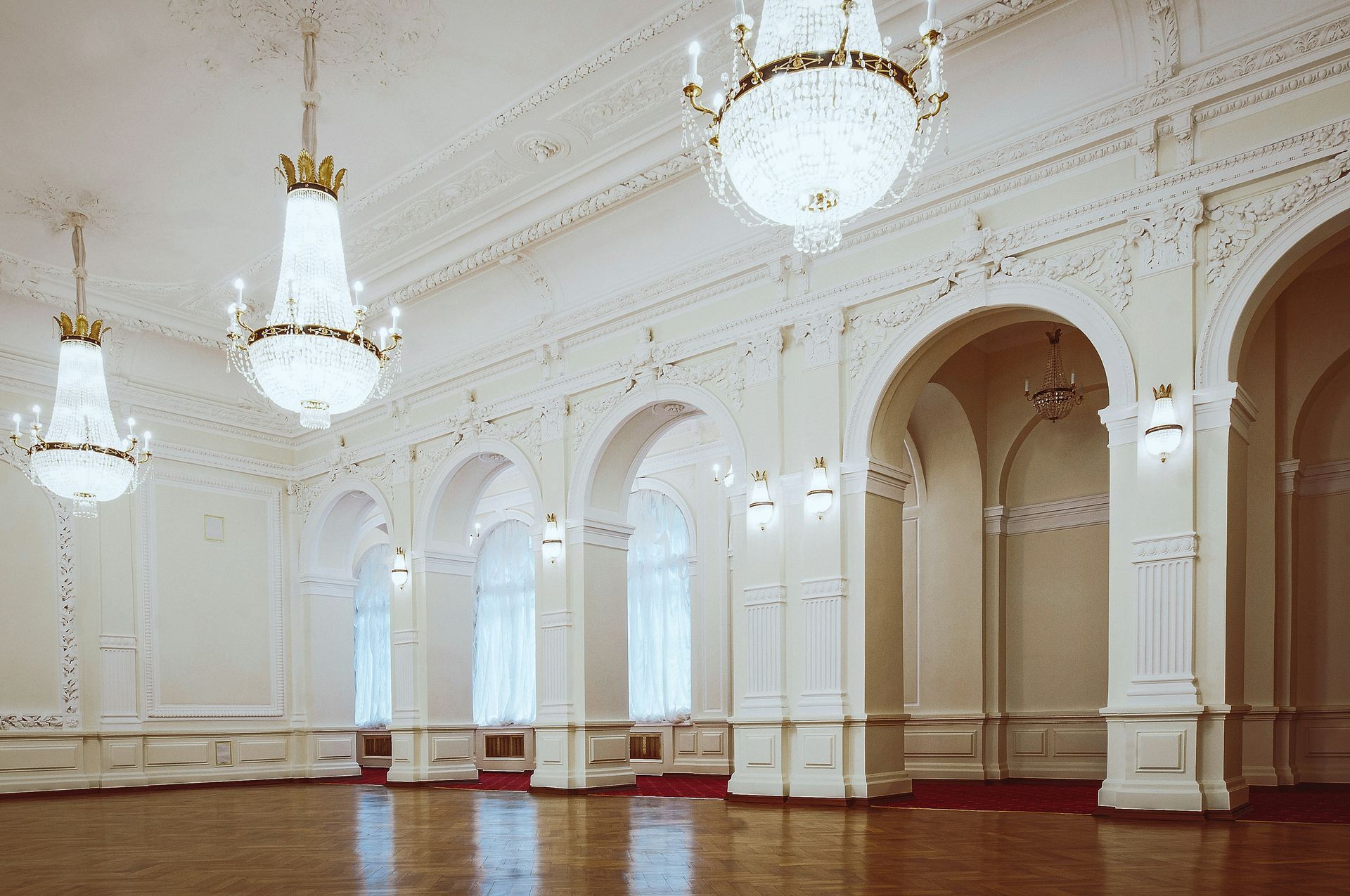 An ornate white ballroom with chandeliers, arched windows, and a wooden floor.