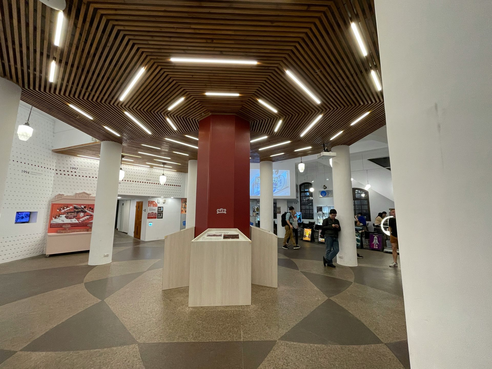 Interior of a building with a red column and a patterned ceiling; people are walking around the space.