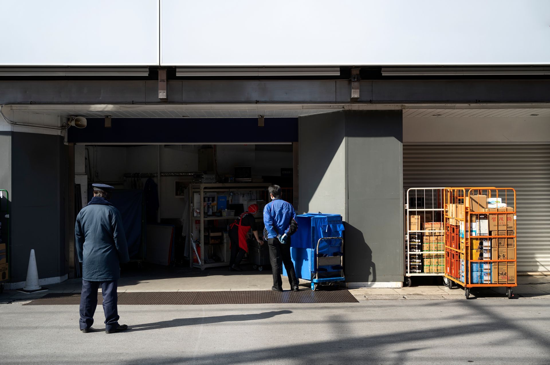 Two people working in a small warehouse; one sorts boxes on shelves, another writes at a desk with a computer.