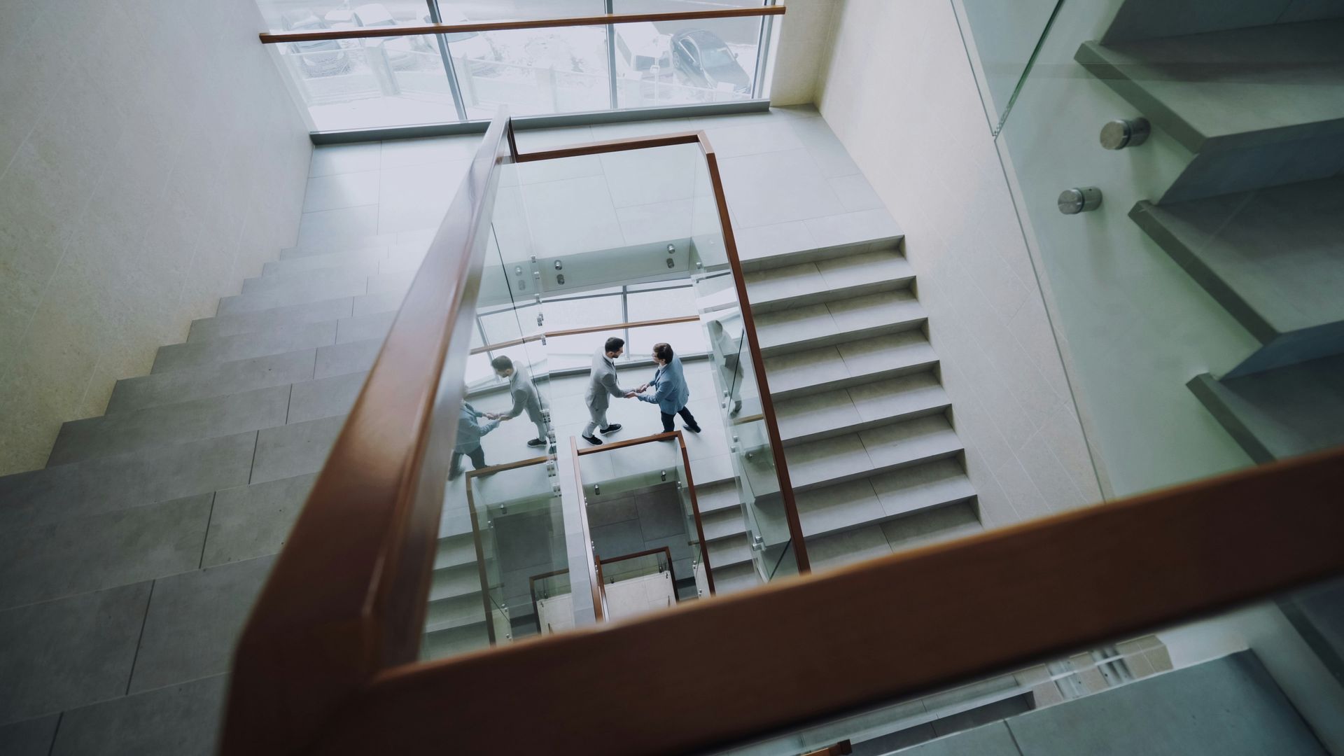 Three people running down stairs, seen from above. Glass walls, light wood banister.