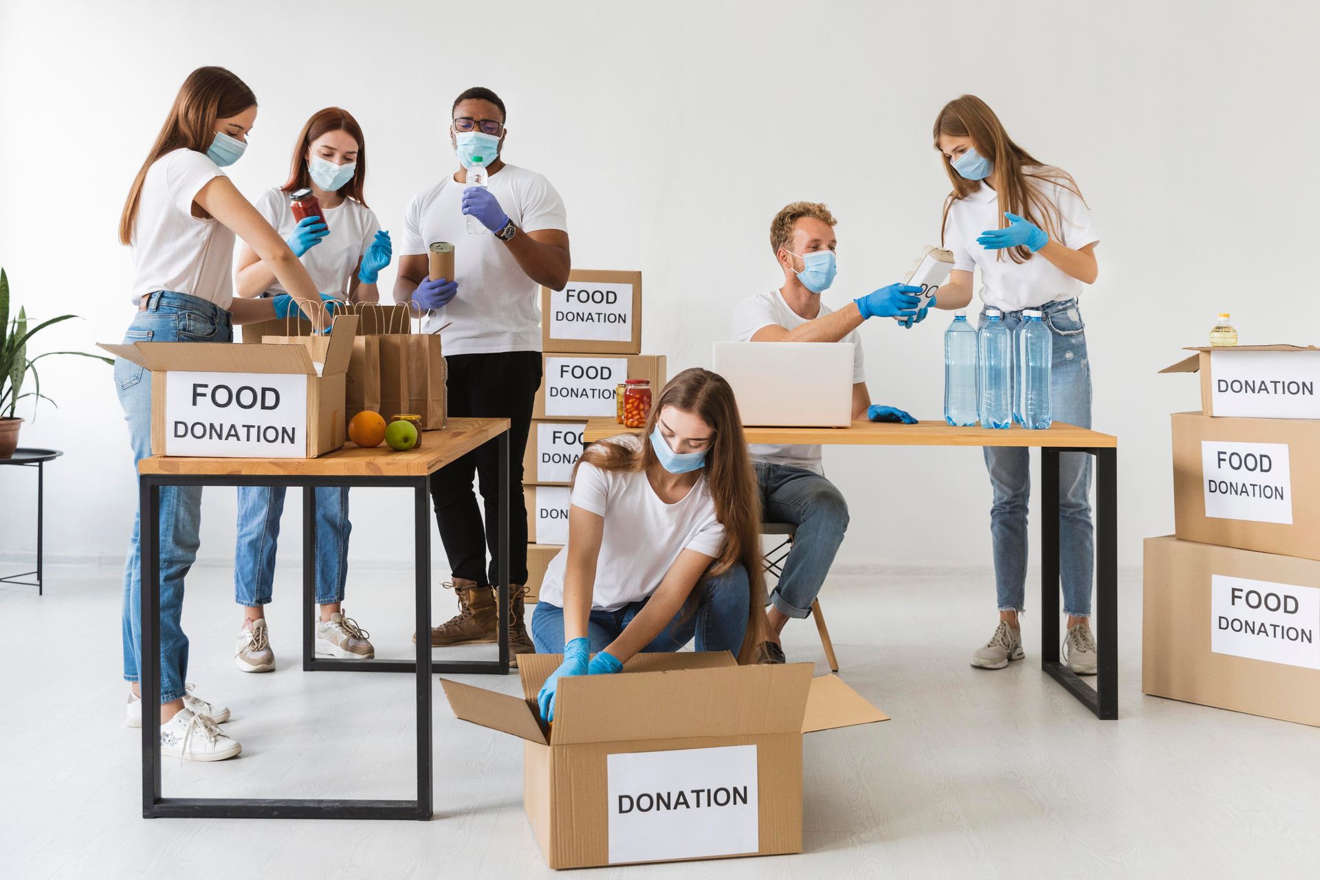Volunteers wearing masks sort food donations into boxes at a bright, indoor workspace.