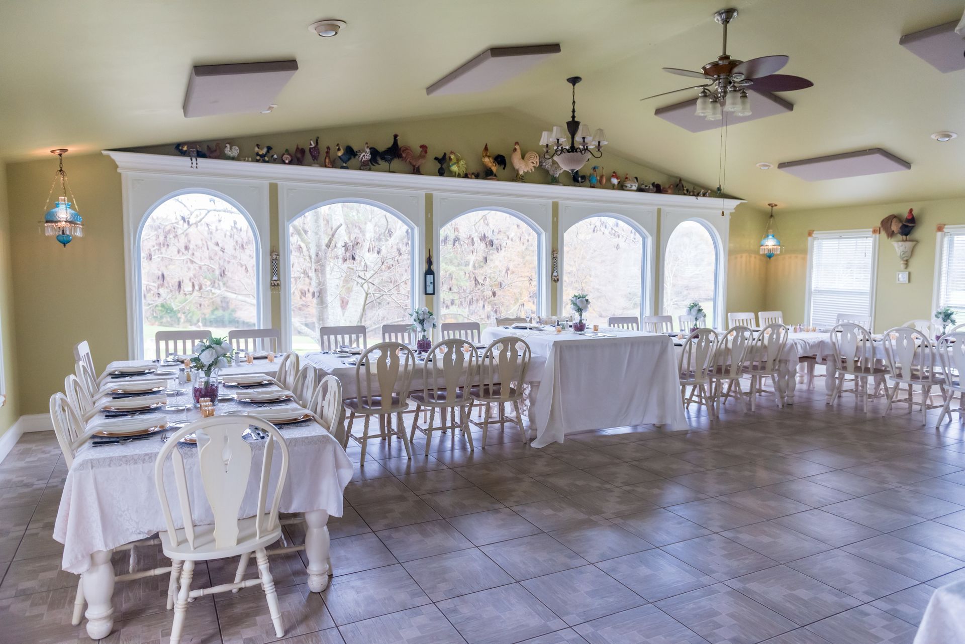 A bright, sunny room with white tables and chairs set for a meal, large windows along one wall.