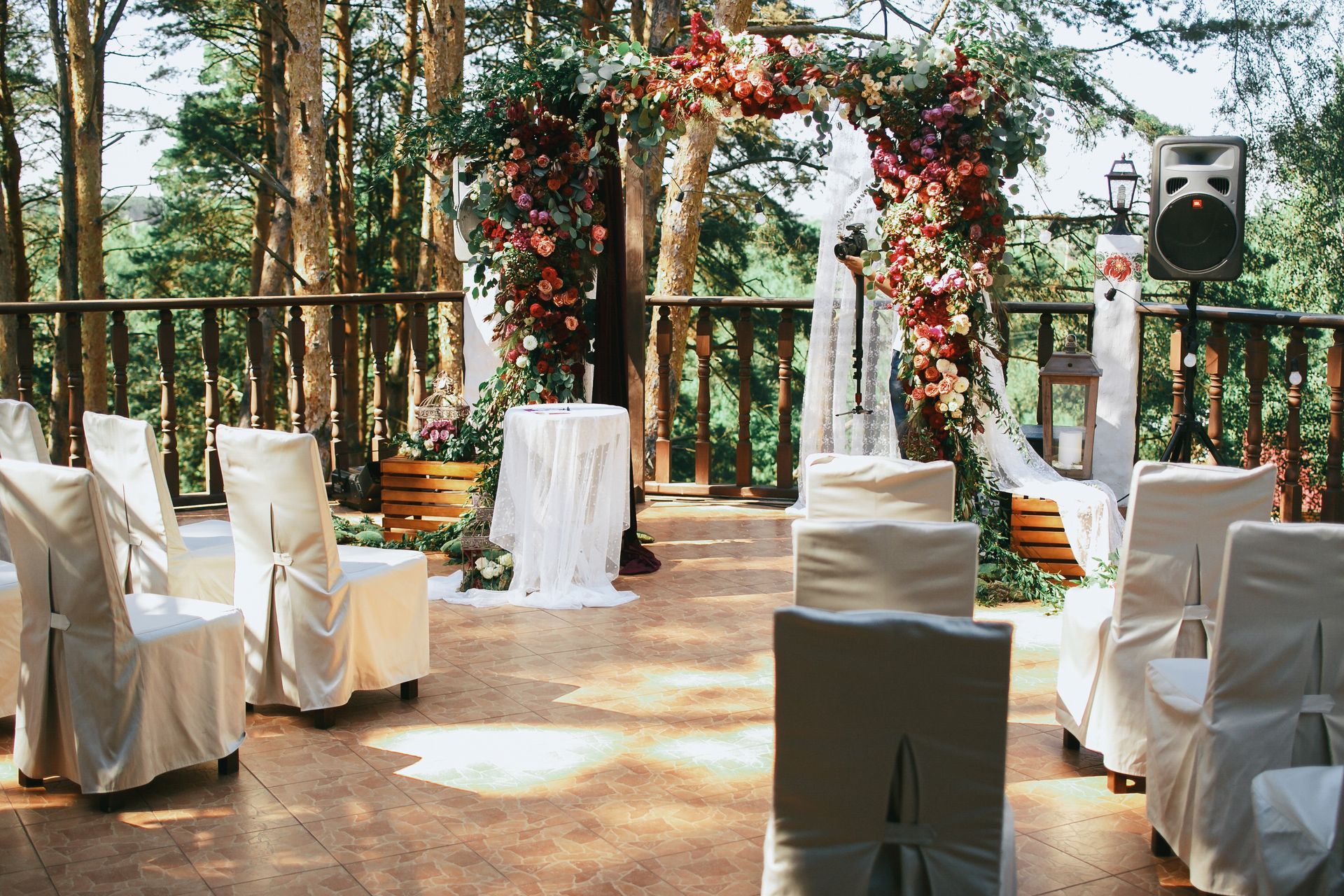 Wedding ceremony setup outdoors with floral arch, draped chairs, and forest backdrop.