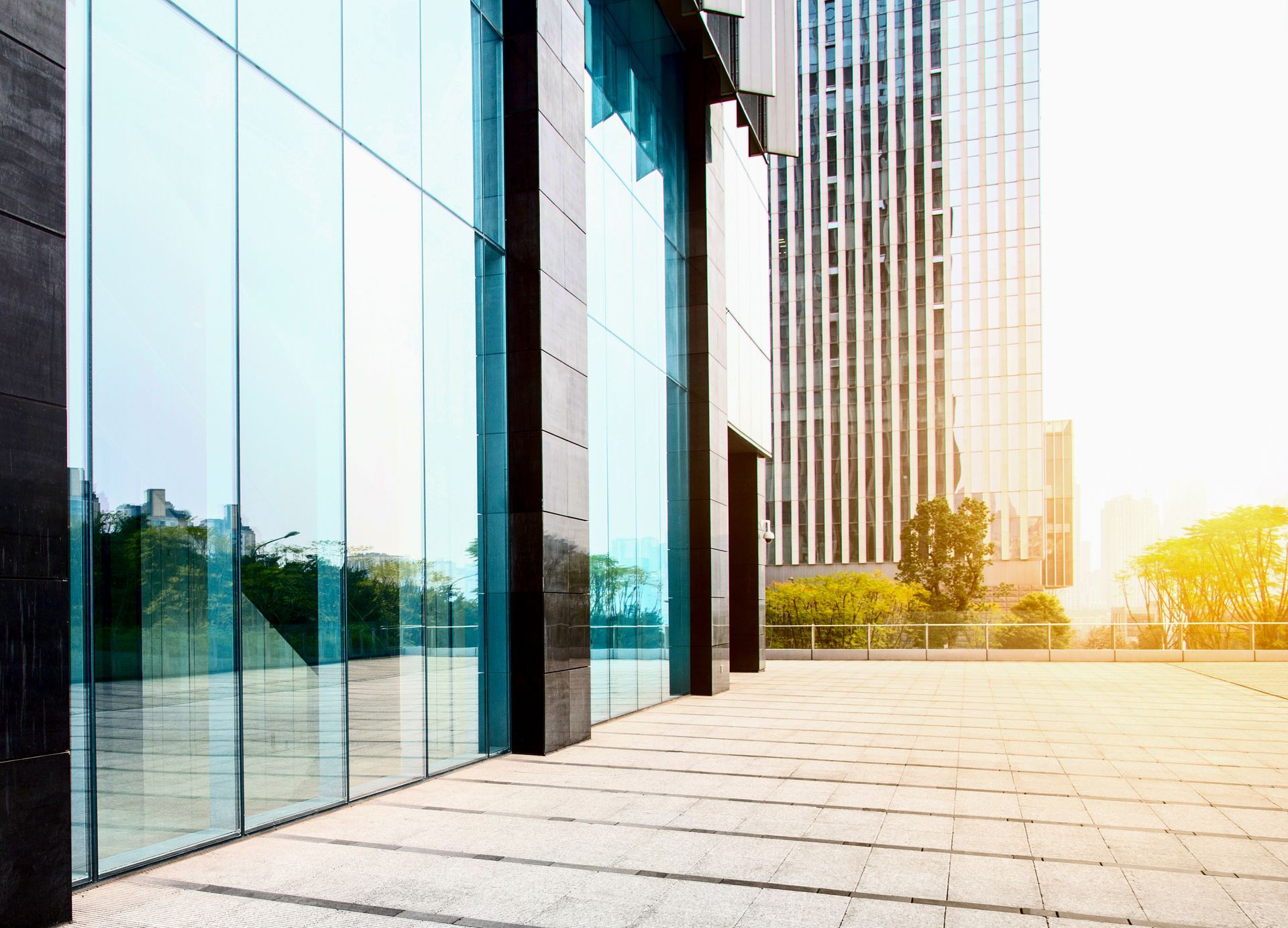Modern glass-walled building facade with a tiled walkway, sunny day, tall skyscraper in background.