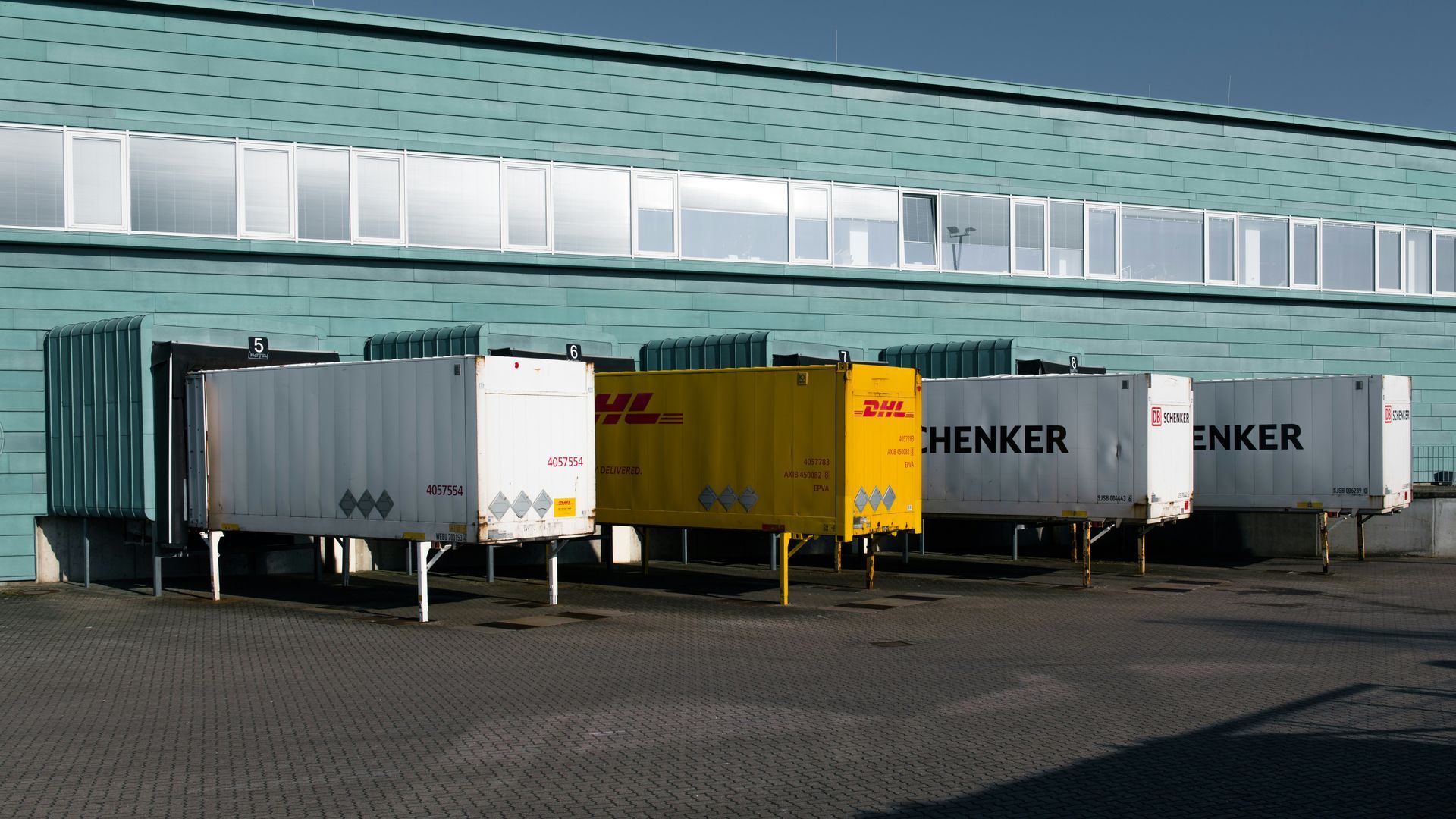 Delivery trucks parked at a warehouse loading dock; DHL, Schenker logos on sides.