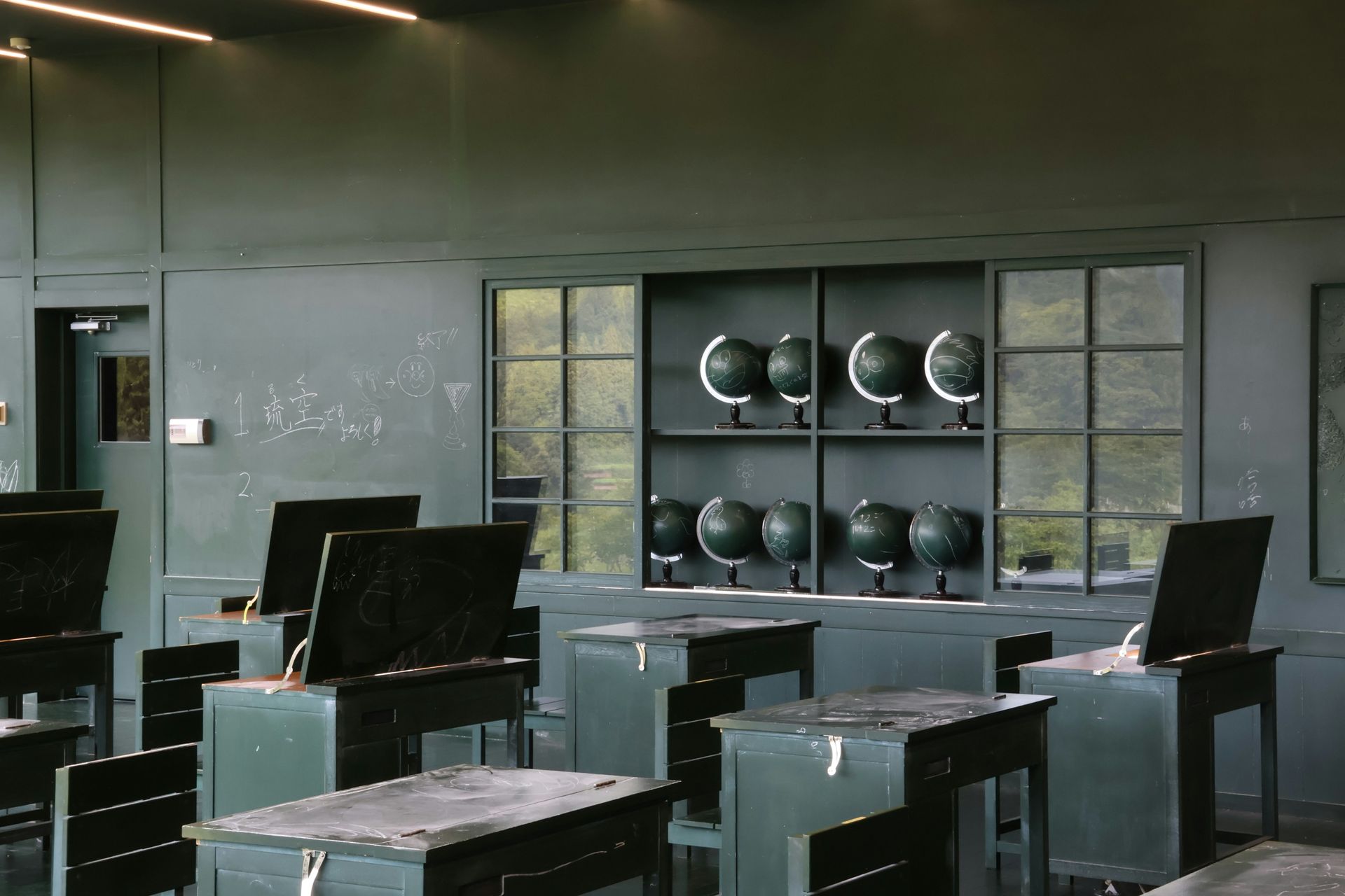Classroom interior, desks facing forward, globes on display. Dark green walls, muted lighting.