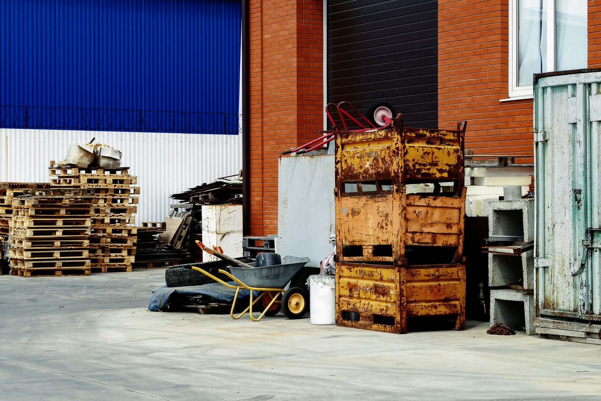 Stack of pallets and rusty containers outside a brick building with a blue door.
