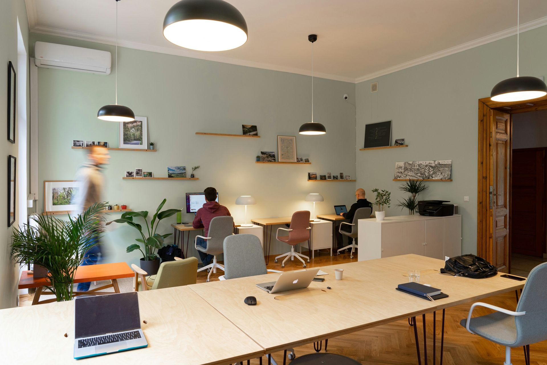 Office with desks, people working, and plants. Light green walls, black pendant lights.