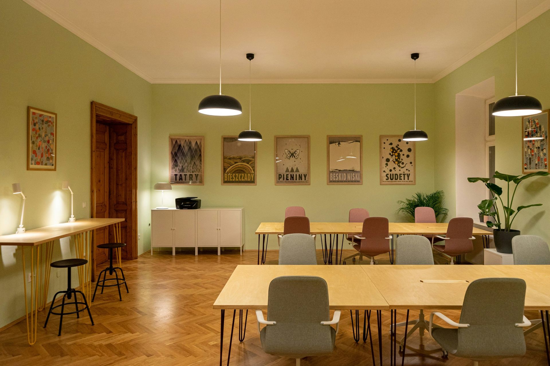 Co-working office space with light wood tables, colorful chairs, green walls, and pendant lights.