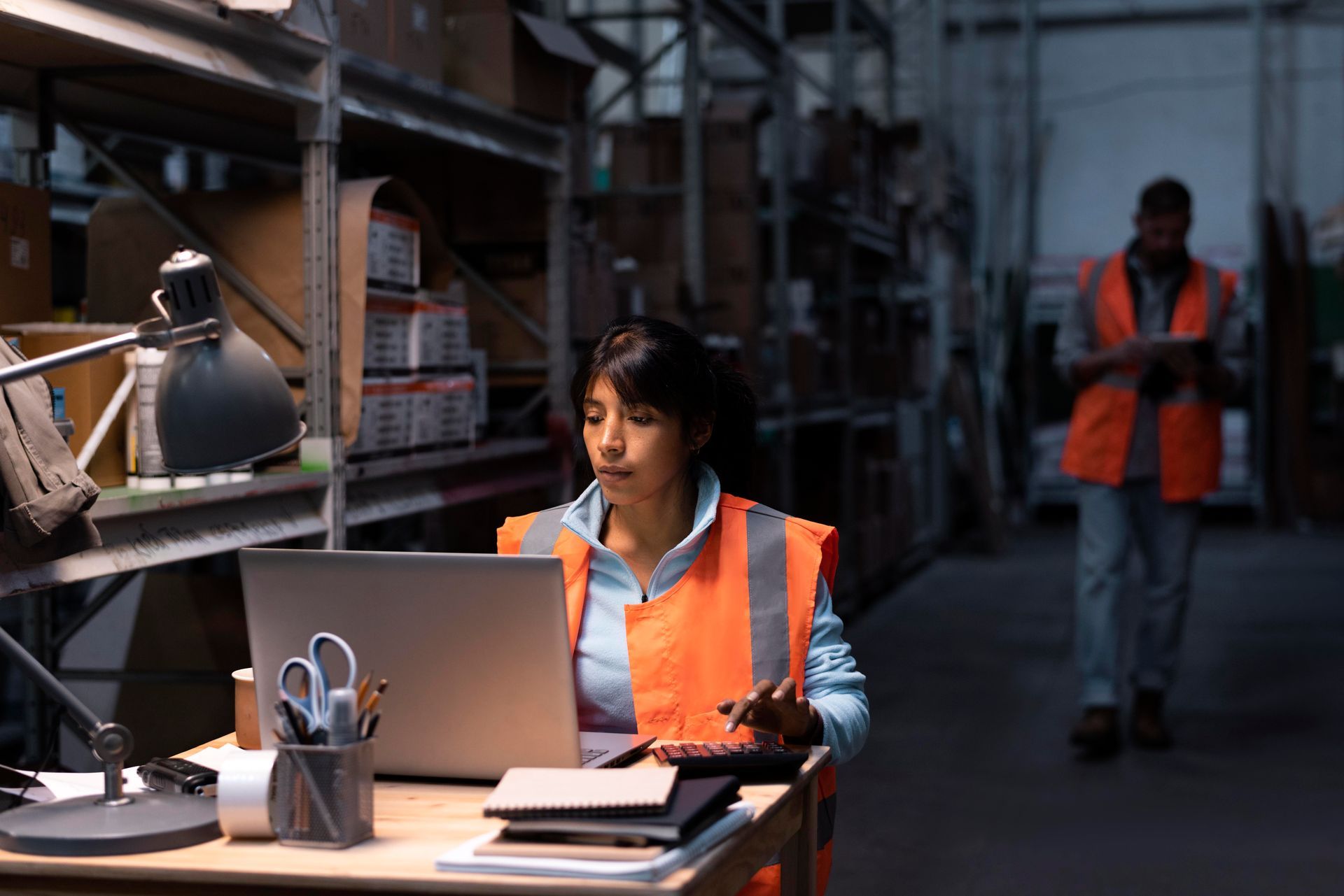 Two people working in a small warehouse; one sorts boxes on shelves, another writes at a desk with a computer.