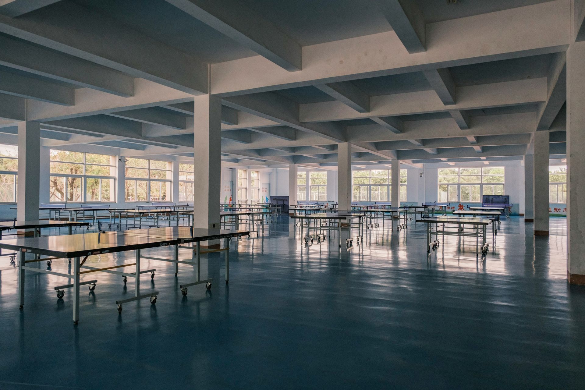 Empty, light-filled cafeteria with rows of tables on a shiny blue floor, windows along the walls, and a structured ceiling.
