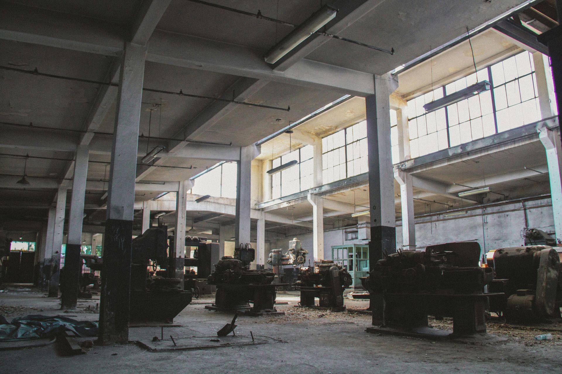 Interior of an abandoned factory with machinery, concrete pillars, and high windows.