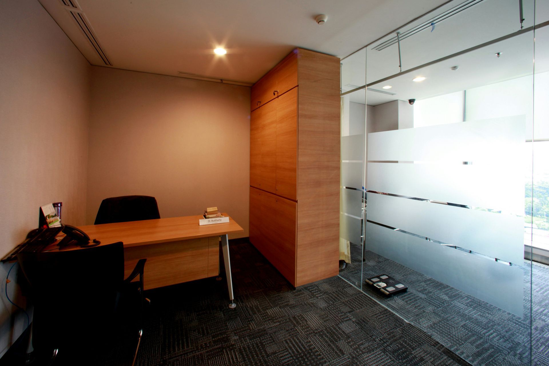 Small office with desk, chair, and wooden cabinet; glass wall with frosted bands.