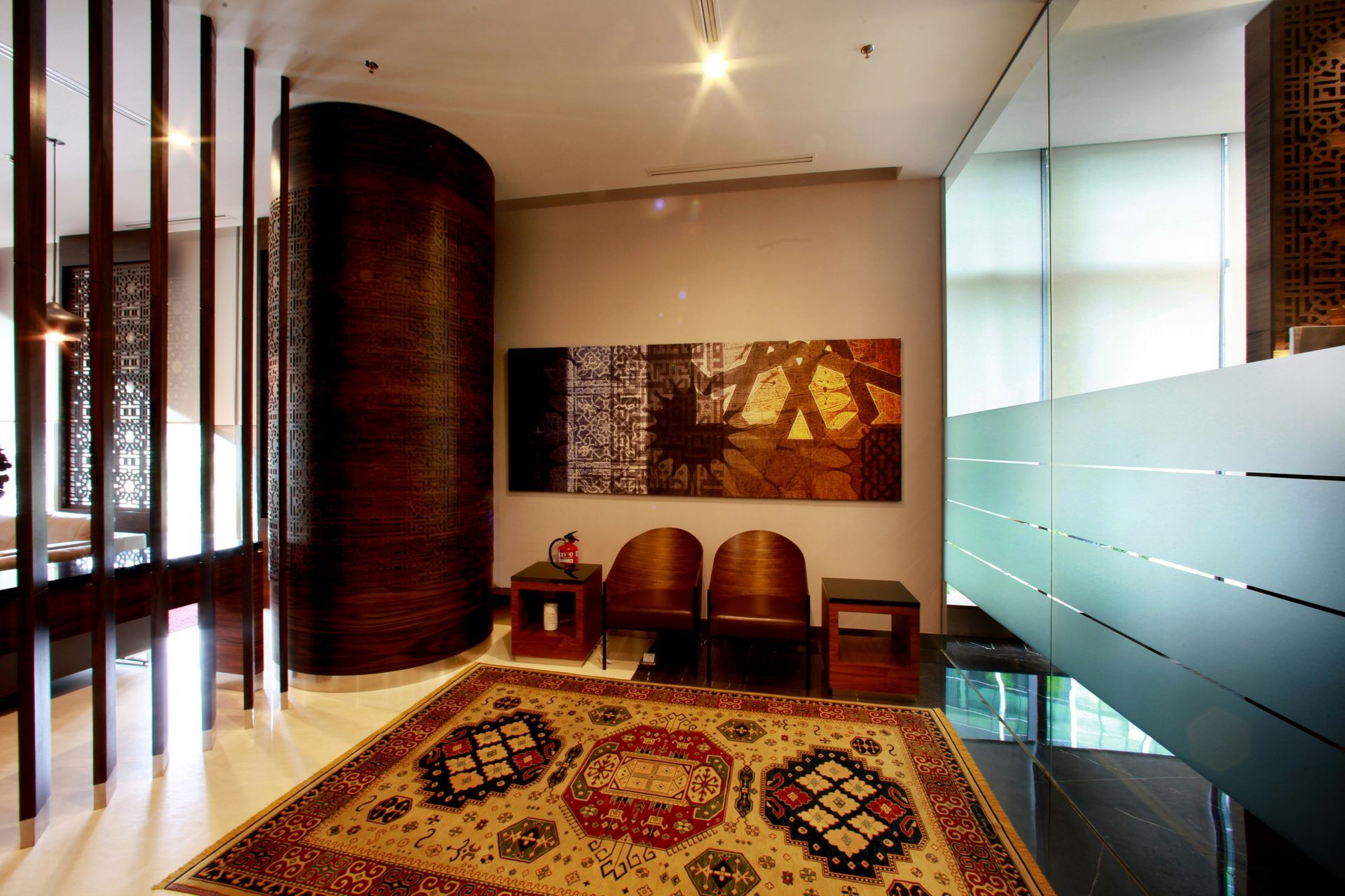 A waiting area featuring wooden accents, a patterned rug, two chairs, and a piece of abstract art.