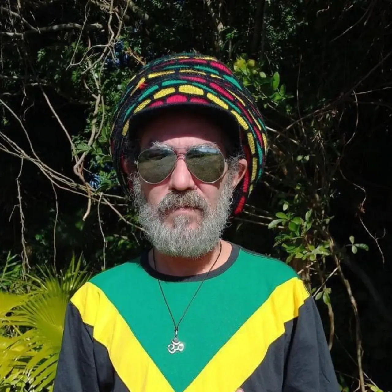 Ras Timbre usando óculos escuros e boina colorida, com camiseta reggae, posando ao ar livre.