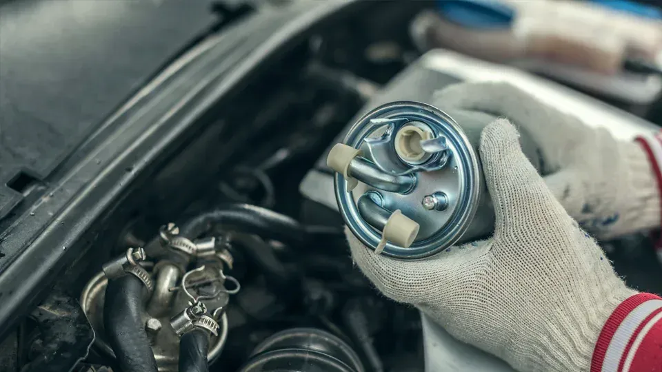A mechanic in gloves holds a new fuel filter near a car engine, preparing to install it.