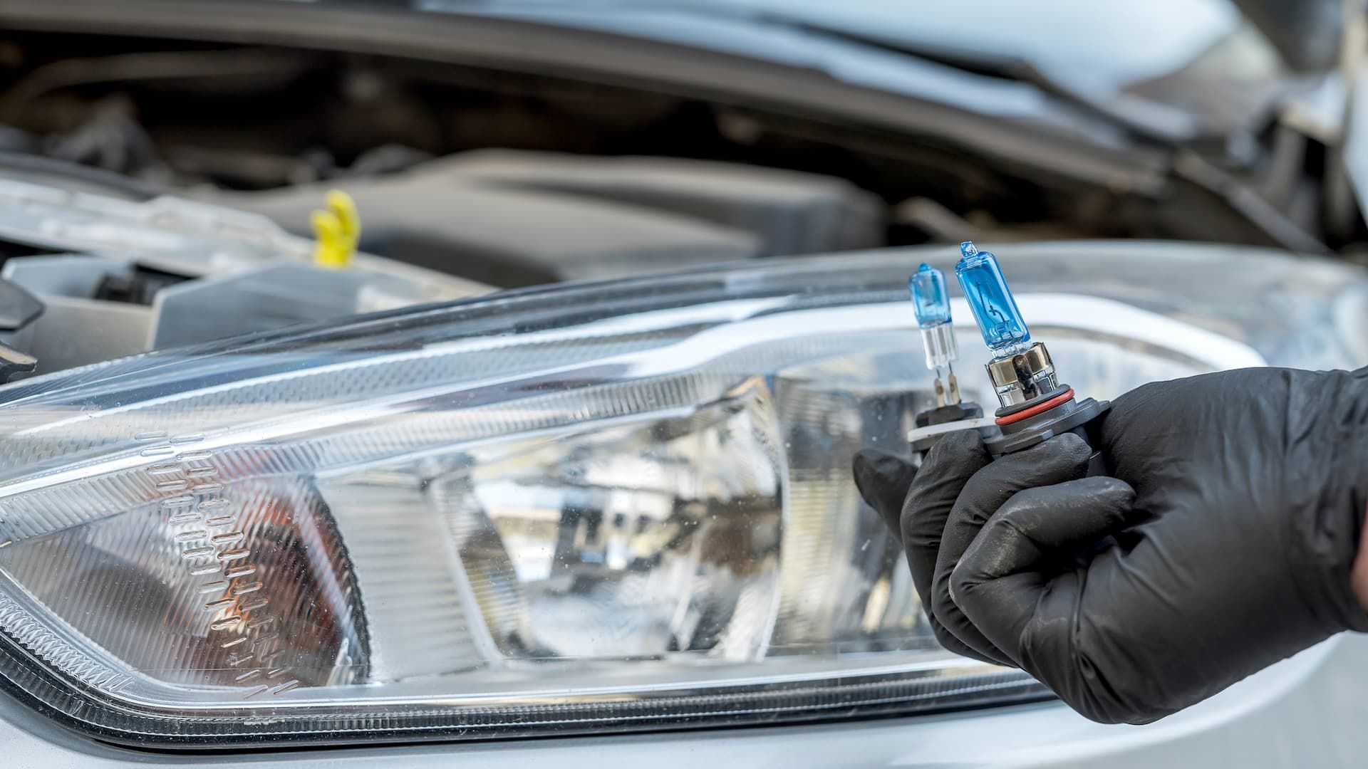 Person in black gloves holding headlight bulbs, replacing one on a car.