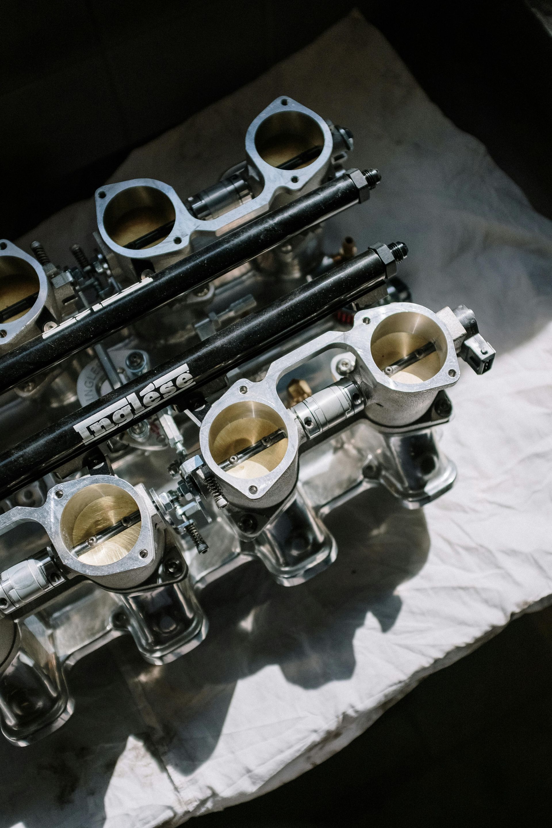 Polished metal intake manifold with four throttle bodies, fuel rails, and open butterflies, on a white surface.