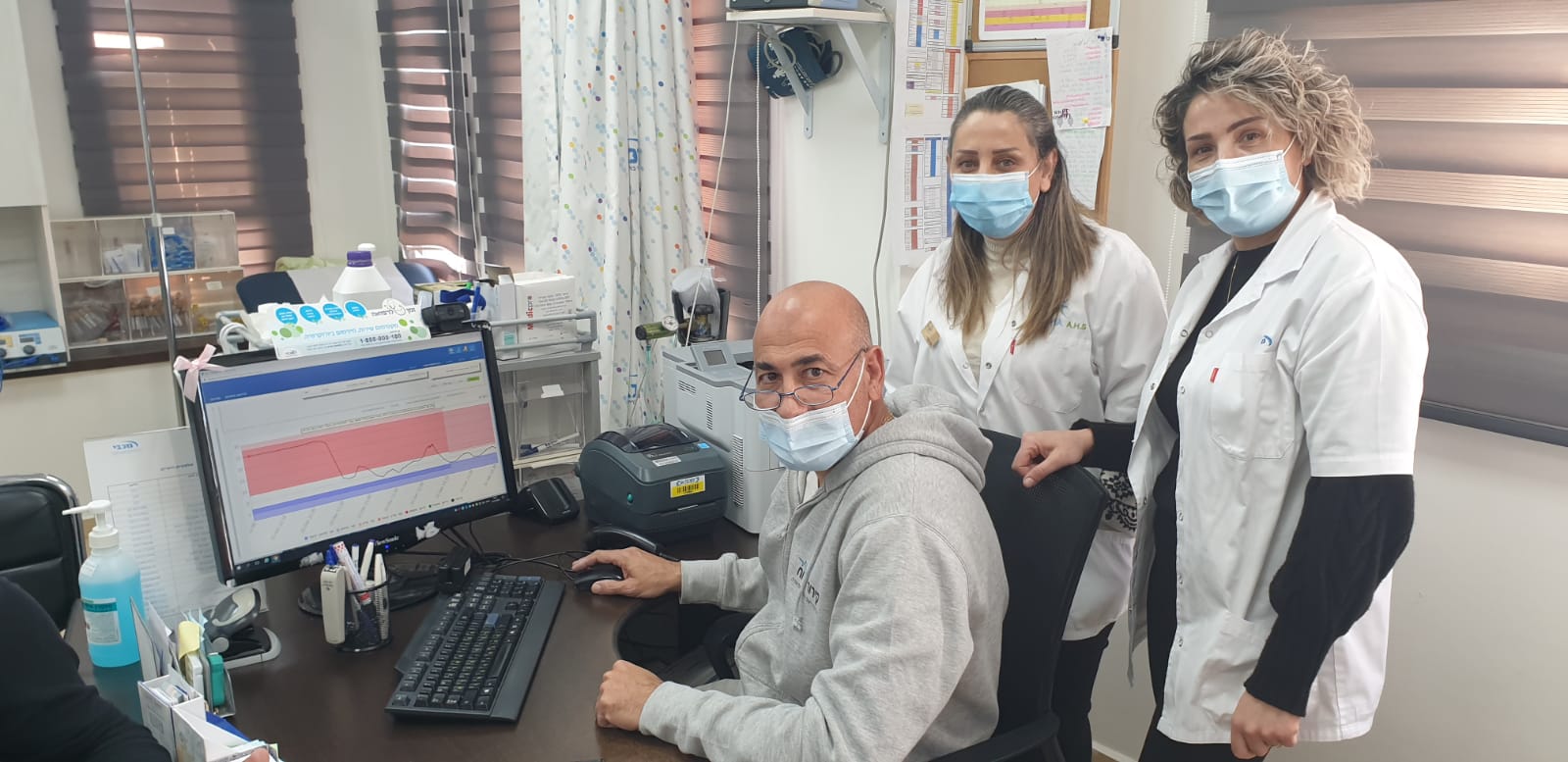 גבר יושב ליד שולחן עם מחשב ושתי אחיות עומדות סביבו.