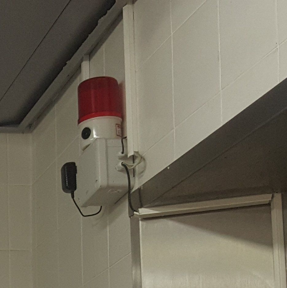 אזעקה אדומה ולבנה מותקנת על קיר מרוצף לבן.