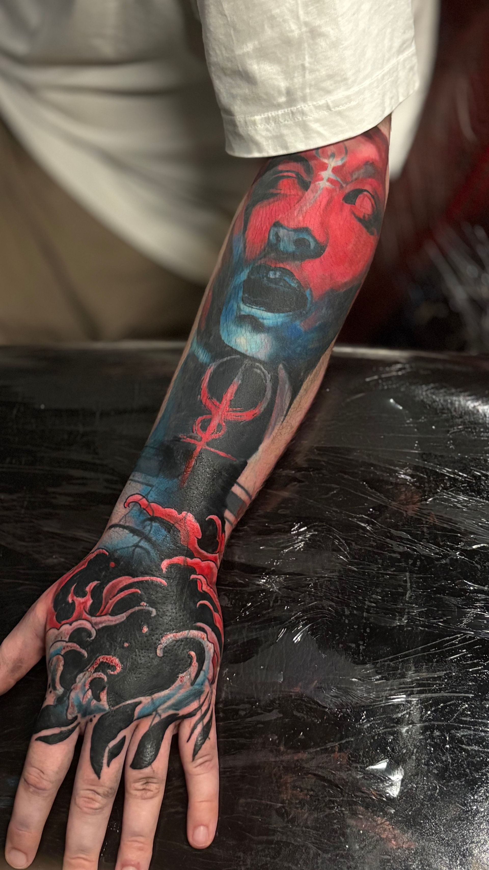 Arm tattoo with a red and blue face, a symbol, and black wave designs on the hand.