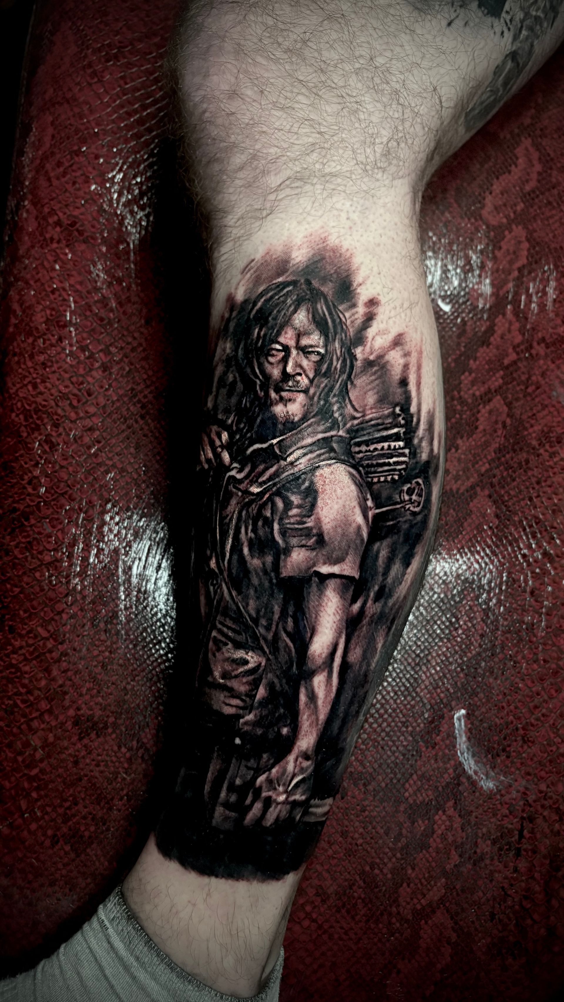 Tattoo of Daryl Dixon from The Walking Dead on a leg. Black and gray portrait, bow and arrow, long hair.