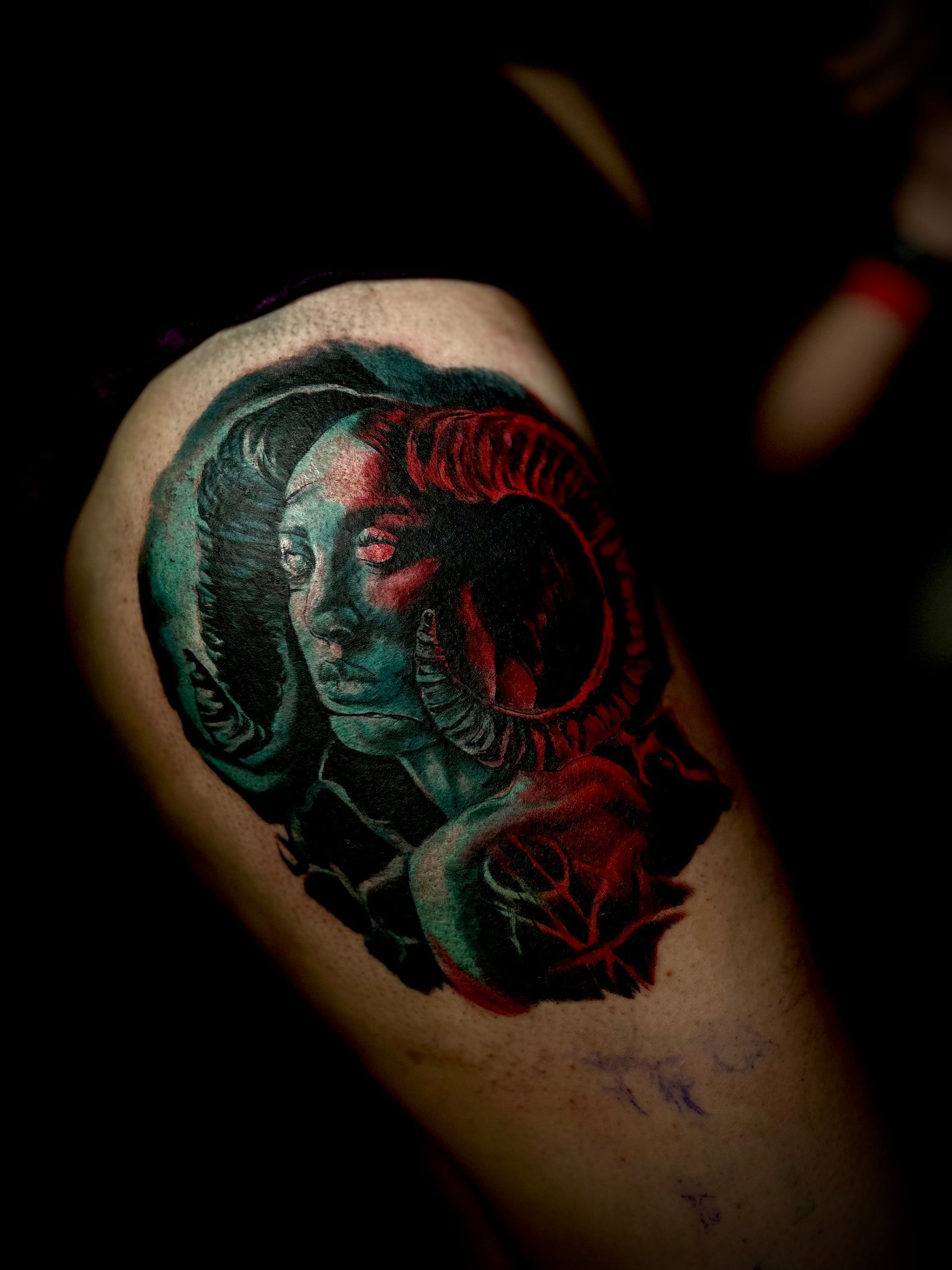 Tattoo of a woman with ram horns; face is teal, horns and parts are red, set against black.