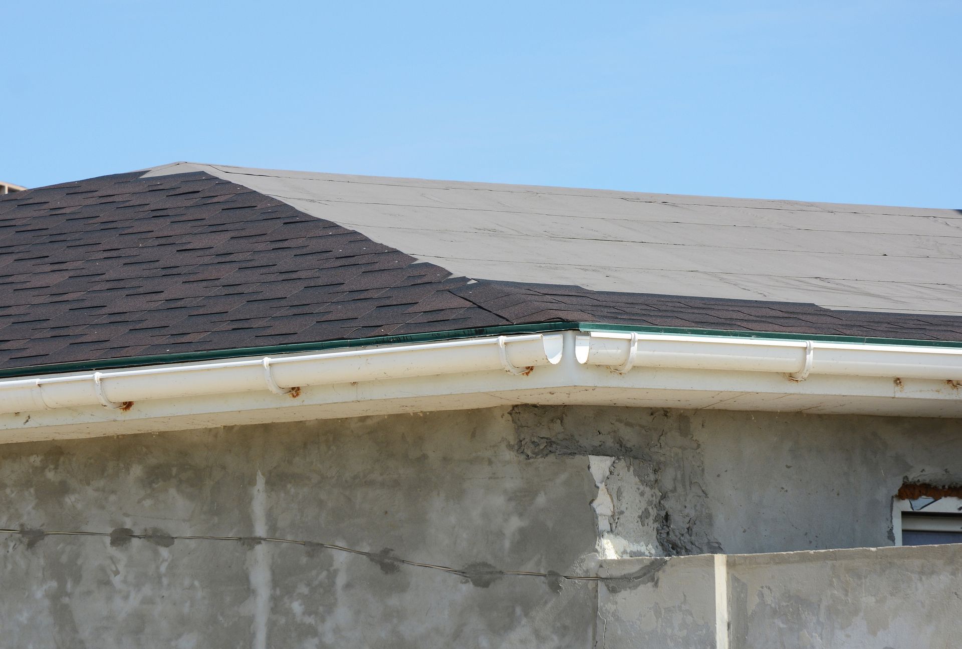 Partially shingled roof, white gutters, and concrete wall against a clear blue sky.