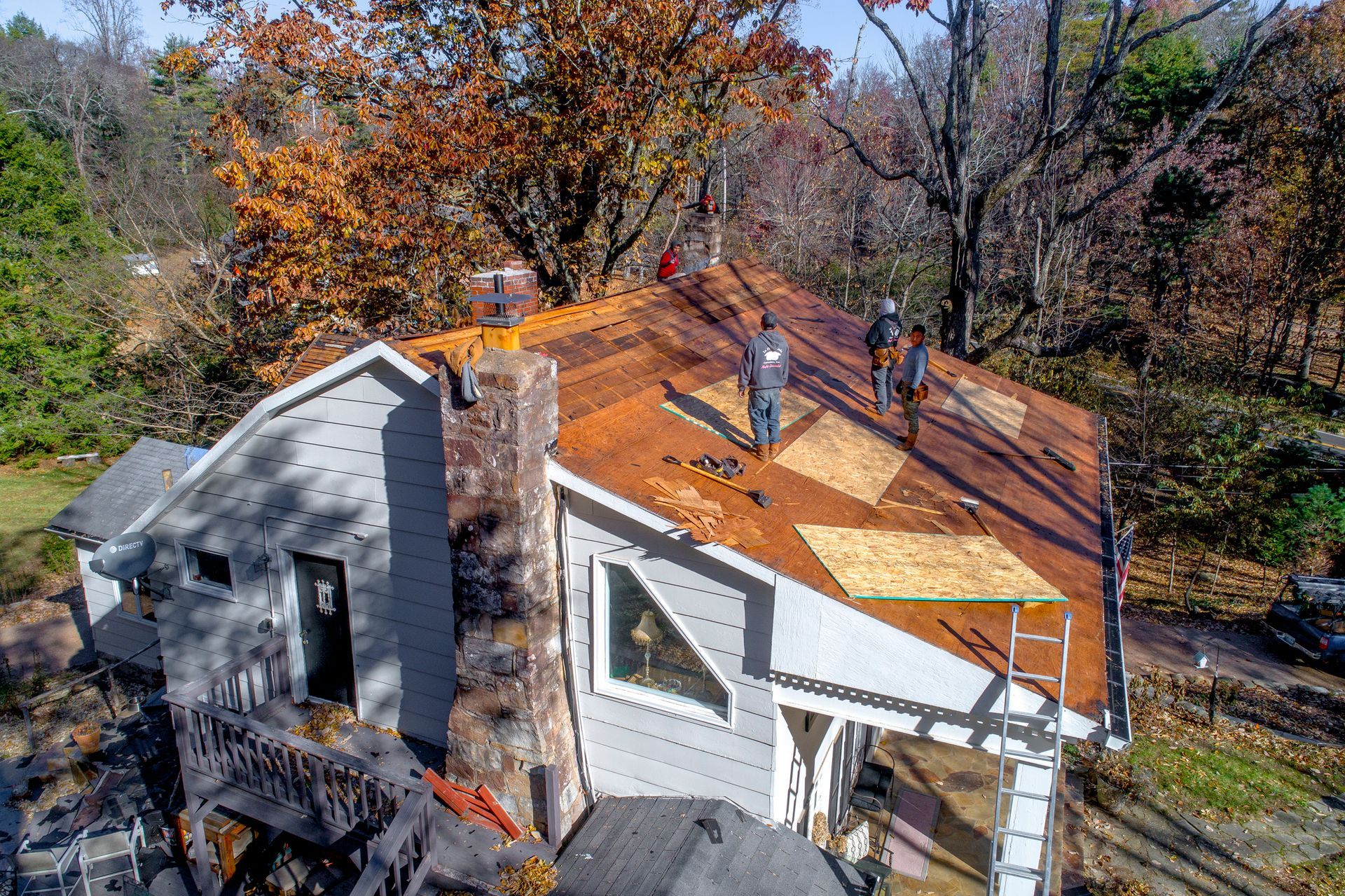 Workers replacing a house roof with tools and a ladder amid autumn trees