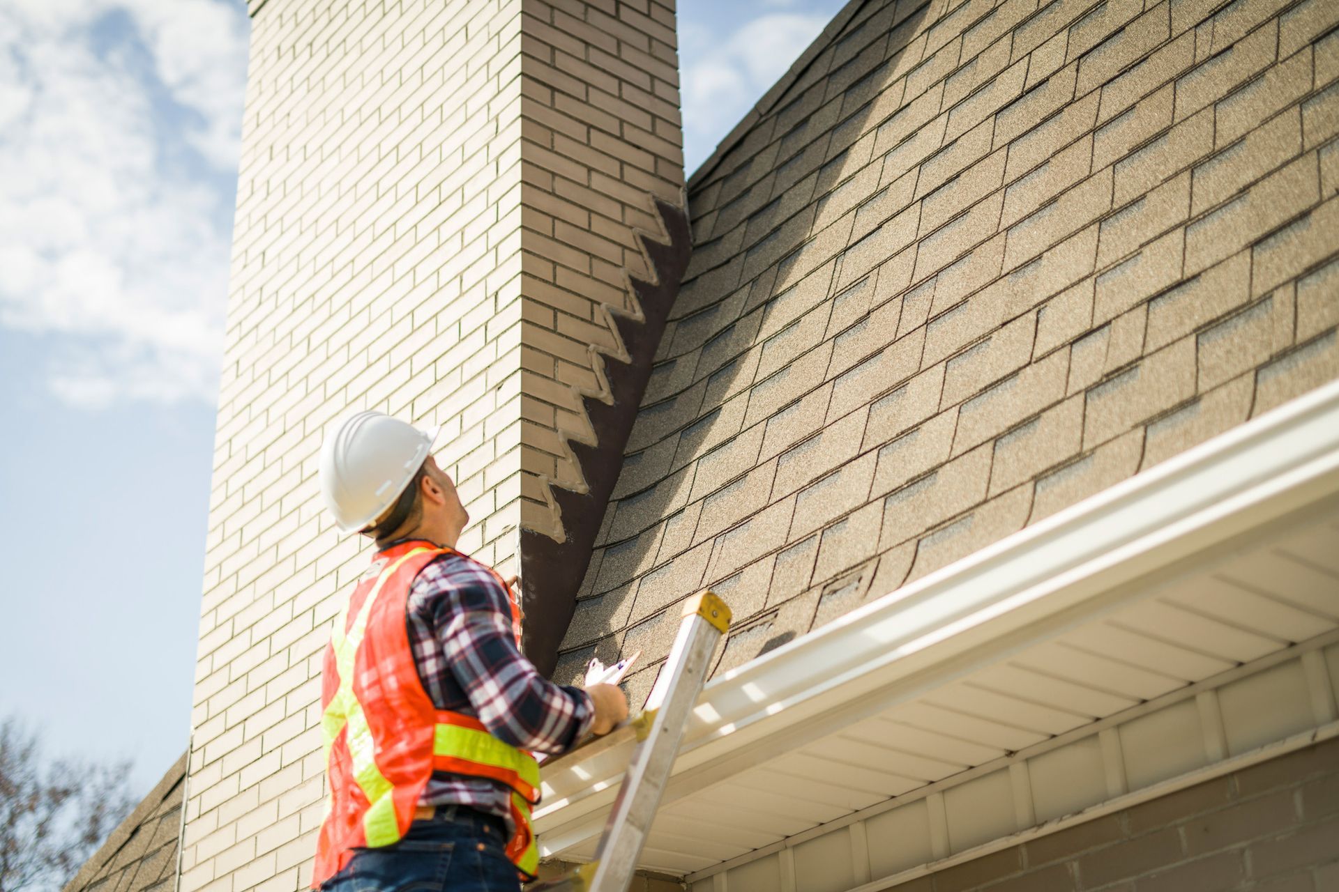 A man with a hard hat is standing on a ladder inspecting a house roof.