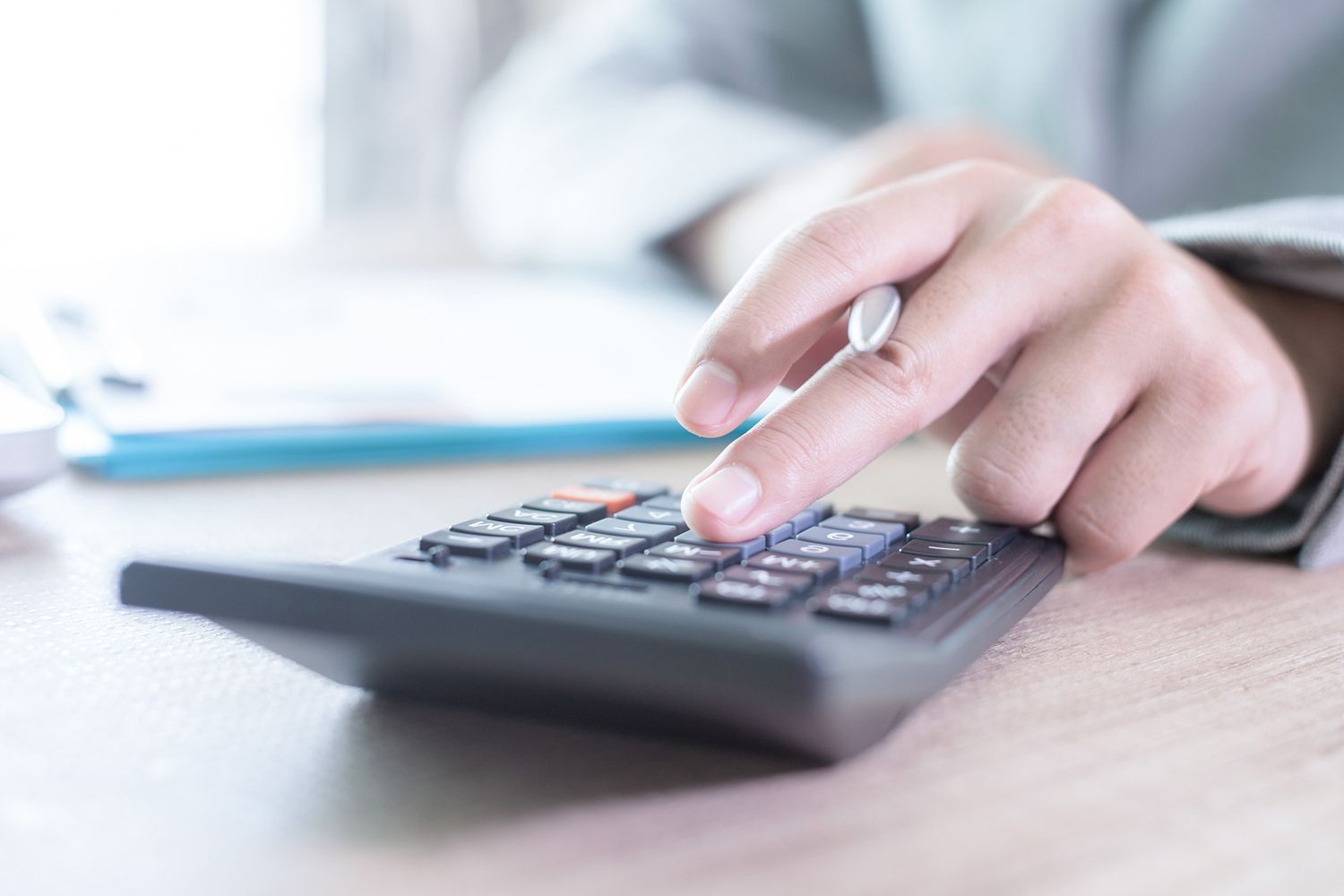 Person's hand with pen calculating on a calculator, blurred office background.