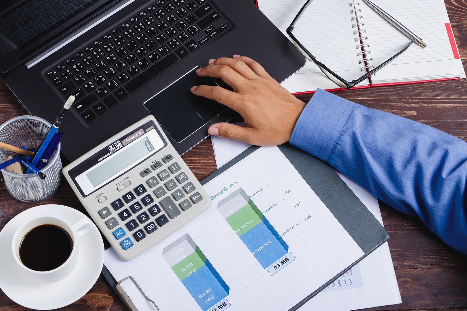 Person working at a desk with a laptop, calculator, and financial charts. Cup of coffee and glasses nearby.
