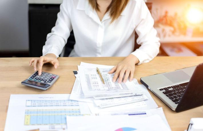 Woman using calculator, reviewing paperwork, laptop nearby. Sunlight shines on desk.