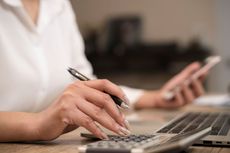 Woman using laptop and smartphone, pen in hand, on a wooden desk.