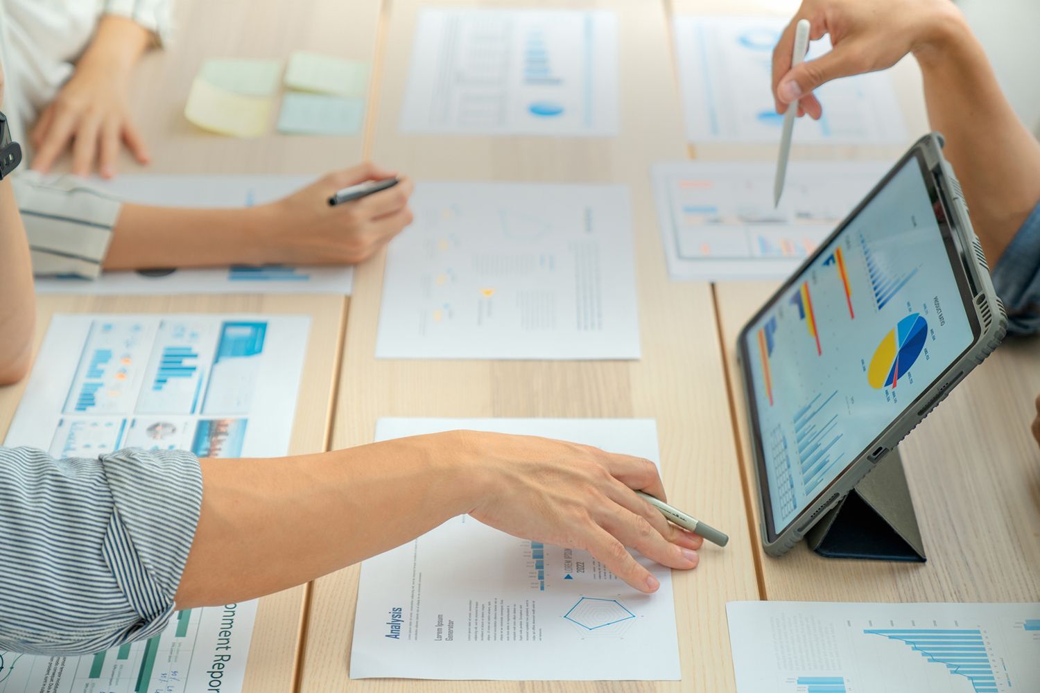 People reviewing charts and graphs on paper and tablet at a wooden table.