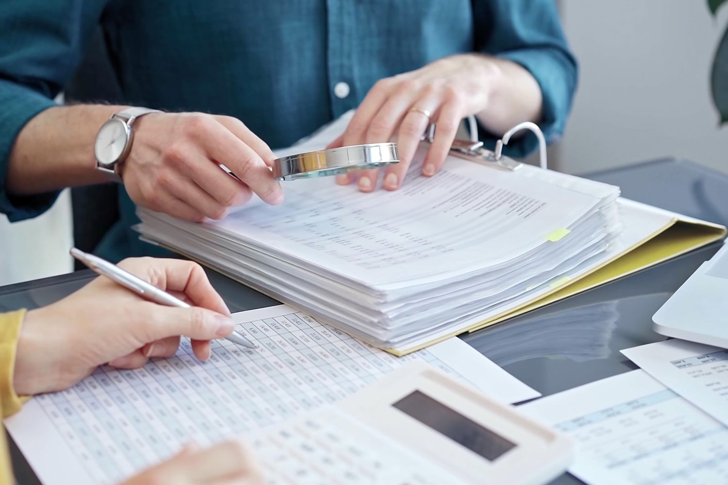 Hands examining documents with a magnifying glass and pen, calculating with a calculator; financial review.