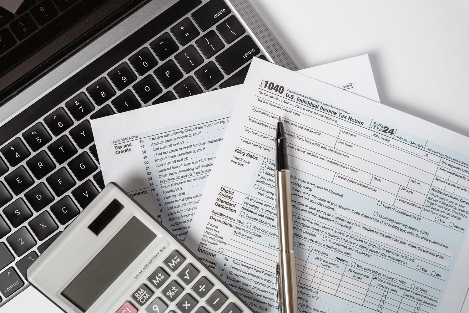 Laptop, tax forms, calculator, and pen on a white surface, suggesting tax preparation.