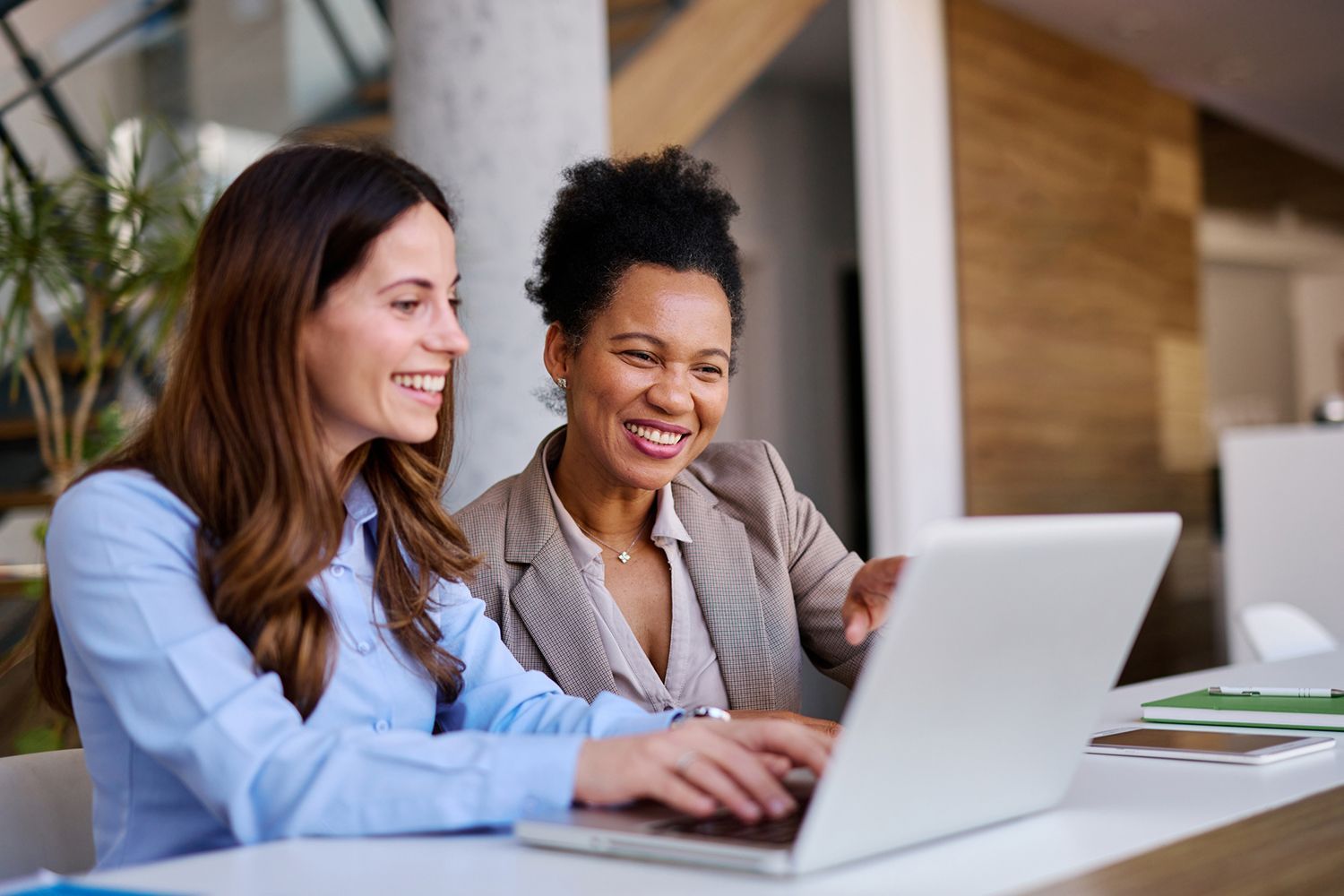 Two women smiling, looking at a laptop in an office, one pointing at the screen.