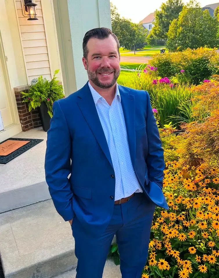 Man in a blue suit smiles, hands in pockets, standing near a house with flowers.