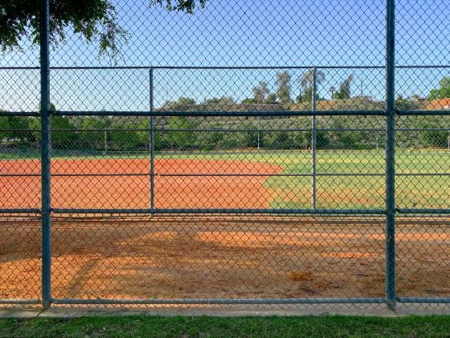 Baseball field behind a chain link fence; red dirt infield and green outfield.