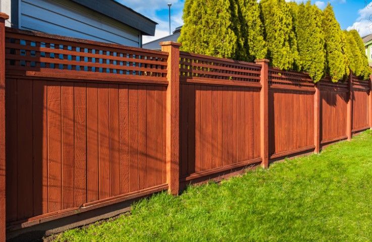 Red wooden fence in a yard with green grass and evergreen trees.