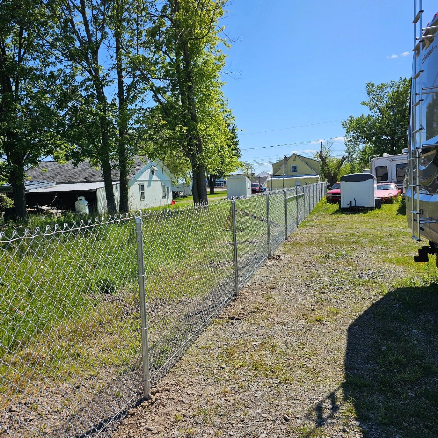 Chain-link fence along a gravel driveway beside trees and houses on a sunny day